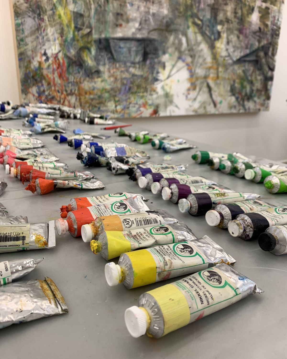 Tubes of oil paint laid out across a grey studio table, a large painting on the wall behind.